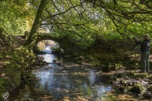 Picturesque Robber's Bridge near Oare - Die malerische Robber's Bridge. Ein Fotograf steht am Ufer eines Flusses und fotografiert eine alte Steinbrücke, die über den Fluss führt. Die Brücke ist von üppigem Grün umgeben, und das Wasser fließt sanft unter ihr hindurch.